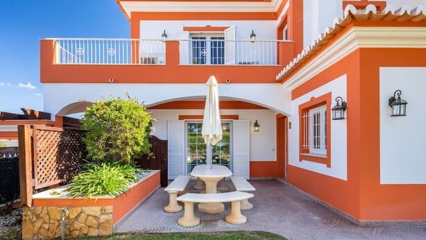 A two-story building with a patio area featuring a table and chairs, a small garden, and a balcony with railings. - Villa Ribeiro II (Albufeira)