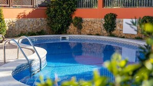 A swimming pool with a stone wall and metal railing, surrounded by greenery and a building with barred windows. - Villa Ribeiro II (Albufeira)