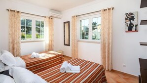 A hotel room with two beds, striped bedding, a large window with curtains, a painting on the wall, and an air conditioning unit. - Villa Ribeiro II (Albufeira)