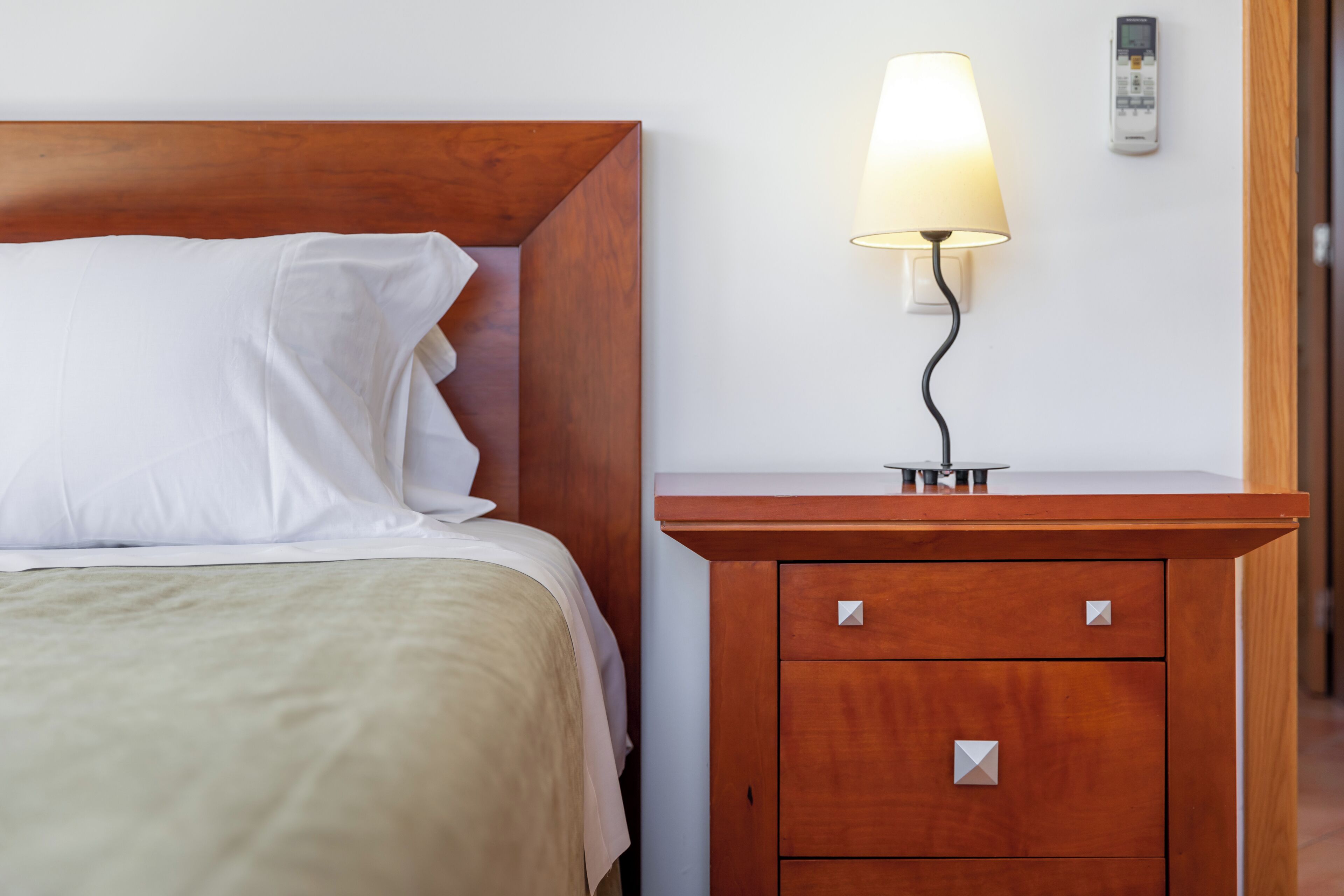 Un lit bien fait, avec du linge de lit blanc, une table de chevet en bois sur laquelle se trouve une lampe, et un téléphone fixé au mur.