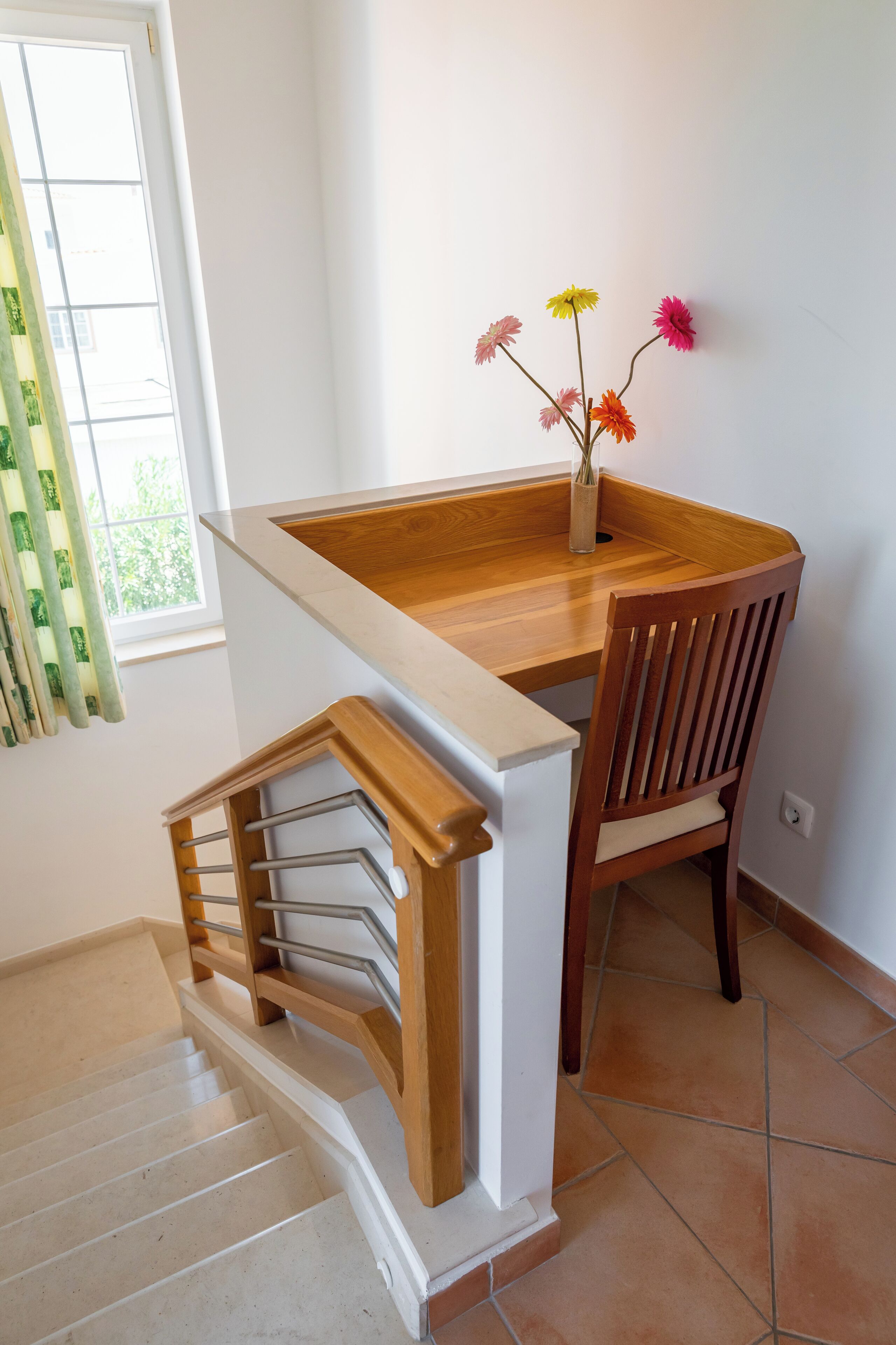A wooden dining table with a vase of flowers, a wooden chair, and a staircase with a handrail.