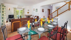 A dining area with a long table set for six, surrounded by red upholstered chairs. A television is visible in the background, and a staircase leads to an upper level. - Villa Ribeiro II (Albufeira)