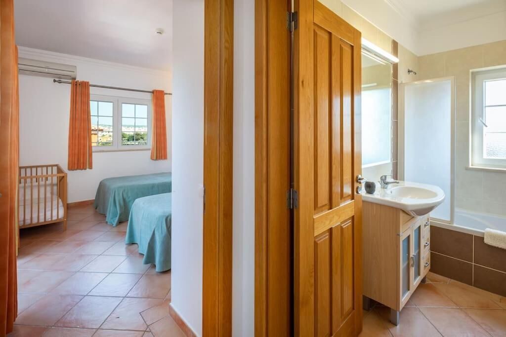 Una habitación con puerta de madera, cama, cuna, ventana con cortinas, lavamanos y espejo.