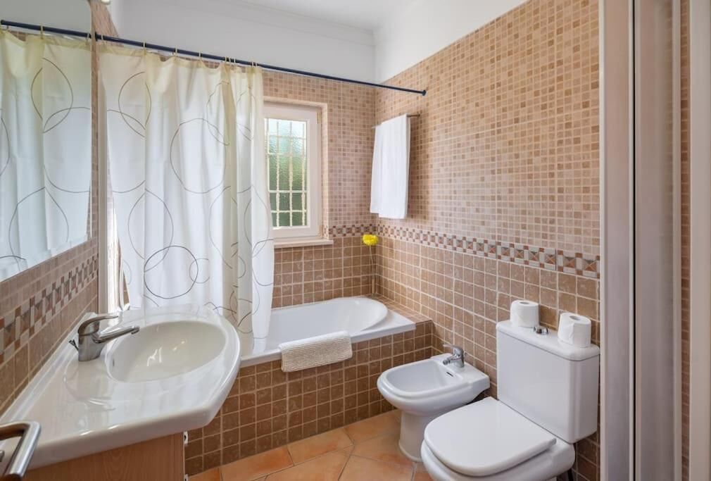 A bathroom with a sink, toilet, bidet, and a window with a curtain.