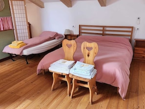 Une chambre avec un lit en bois, une table de chevet, une chaise avec des serviettes, et un futon en arrière-plan.