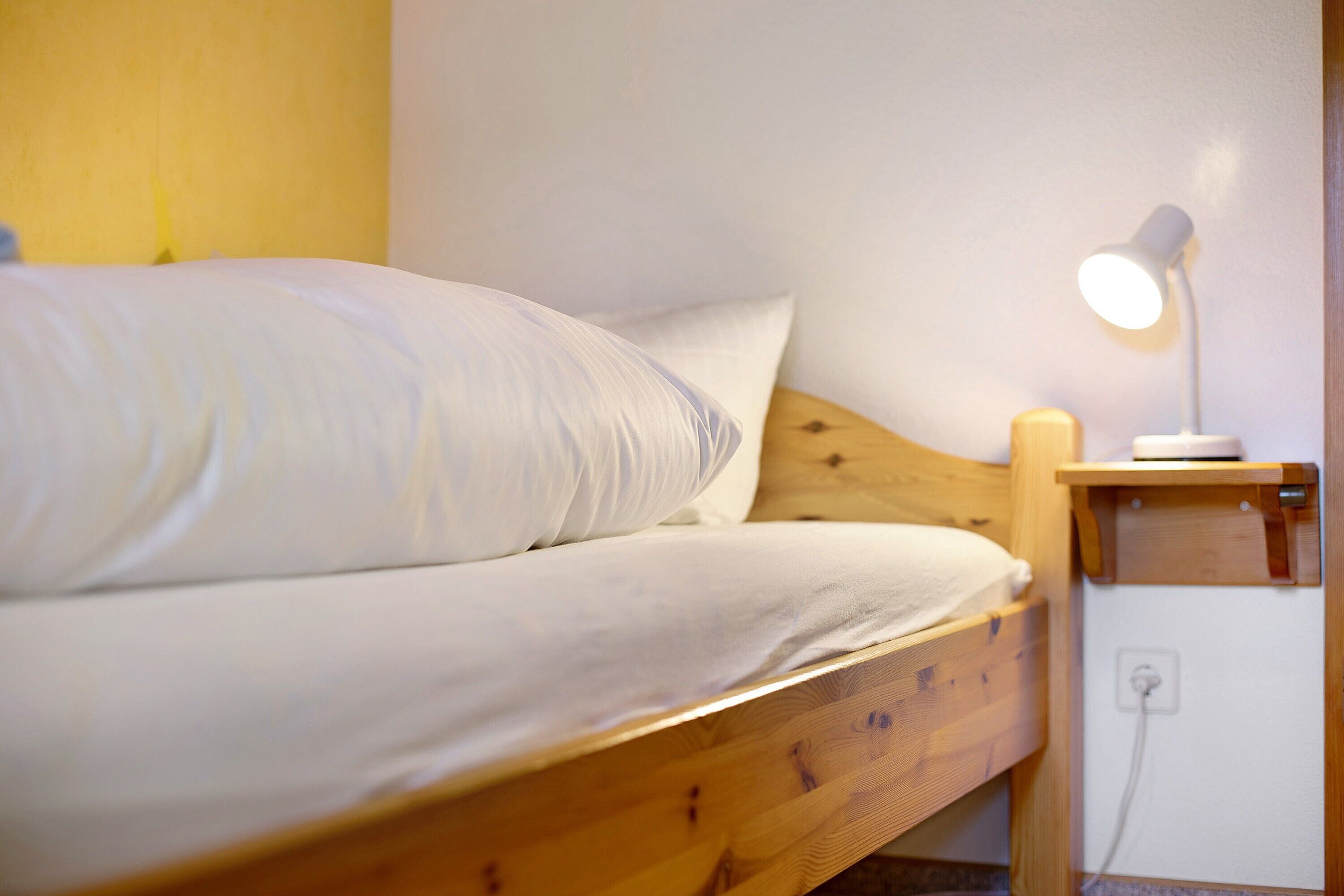 A single bed with a wooden frame, a white mattress, and a bedside table with a lamp.