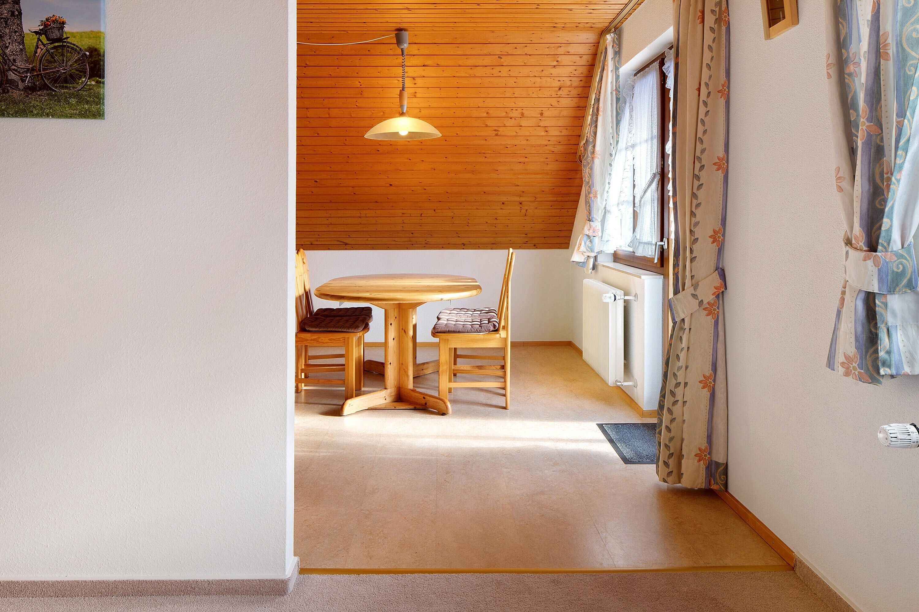Et træbord med stole, en hængende lysarmatur og et vindue med gardiner.
