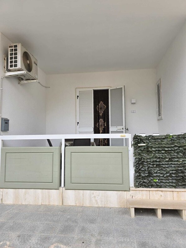 A balcony with a white air conditioning unit, a closed door with horizontal blinds, and a green wall with a leafy plant. - Two-room Seafront Apartment - Torre Canne (Fasano) (Torre Canne)