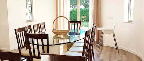 A dining area with a glass table surrounded by wooden chairs, a wicker basket, and a view of greenery through a large window.