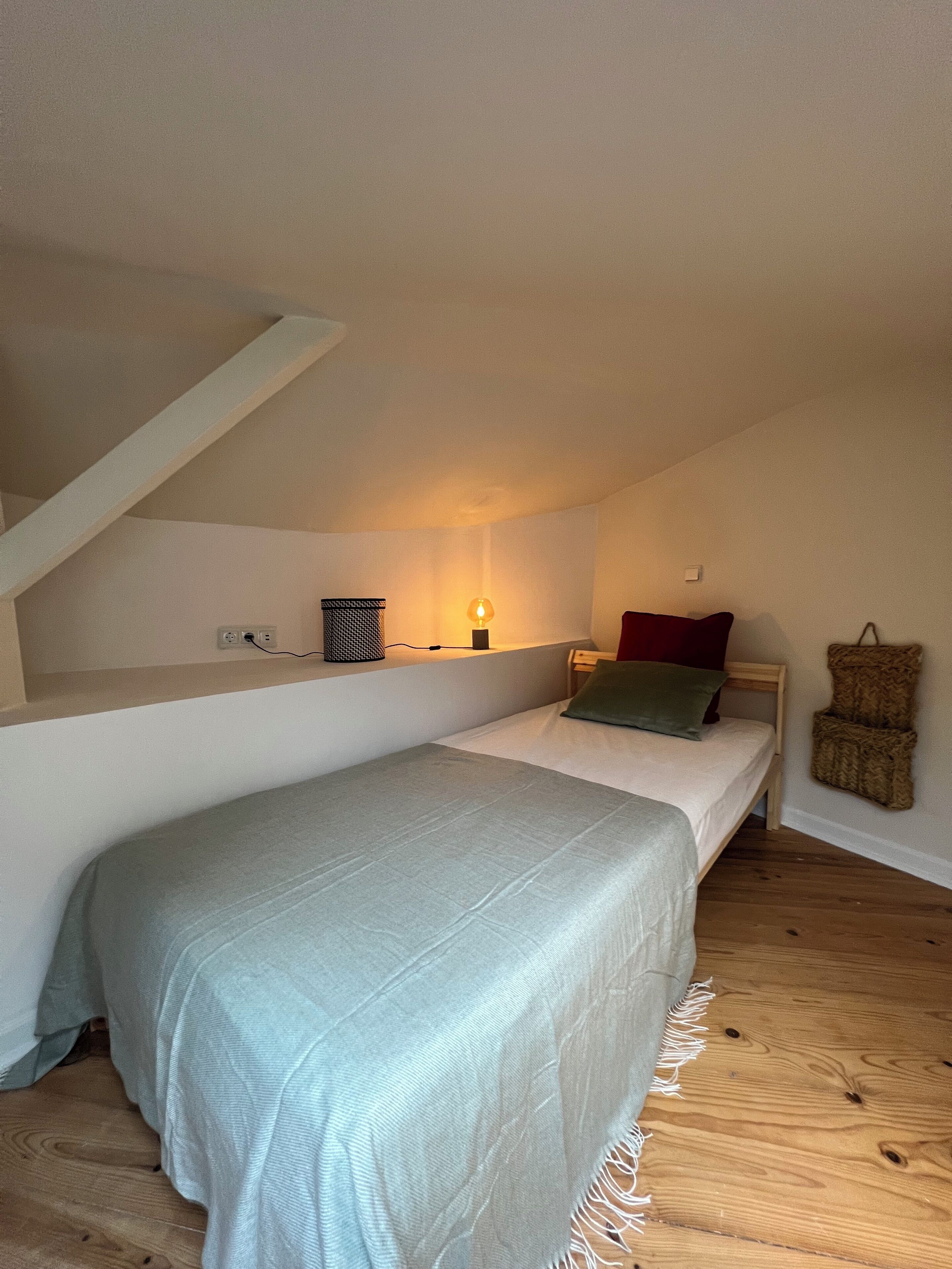 Ein kleines, gemütliches Schlafzimmer mit einem Einzelbett, einem Nachttisch, einer Lampe und einer hängenden Körbchenampel.
