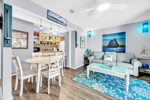 A modern living room with a gray sofa, a coffee table, and a dining area with a white table and chairs. - 🏖️Walk to Beach | 2 Pools | Great Amenities | Endless Summer E10🏖️ (Panama City Beach)