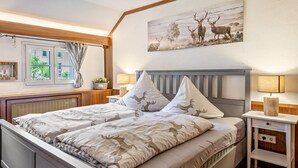 Une chambre à coucher comprenant un lit, deux tables de chevet, une fenêtre avec des rideaux, un tableau représentant des cerfs accroché au mur et un radiateur.