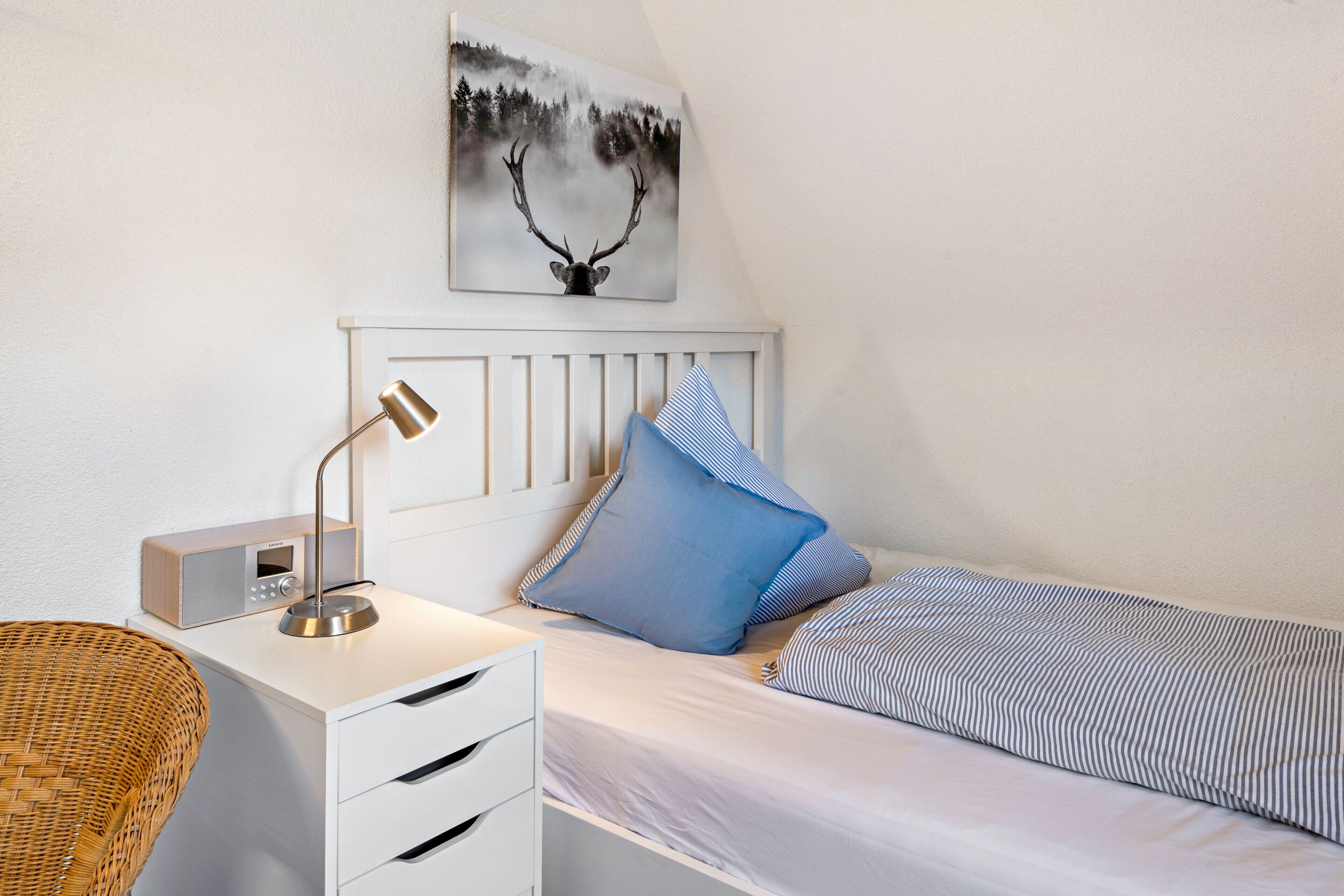 Ein Schlafzimmer mit einem Bett, einem Nachttisch, einer Lampe, einem Radio und einem Bild von einem Reh an der Wand.
