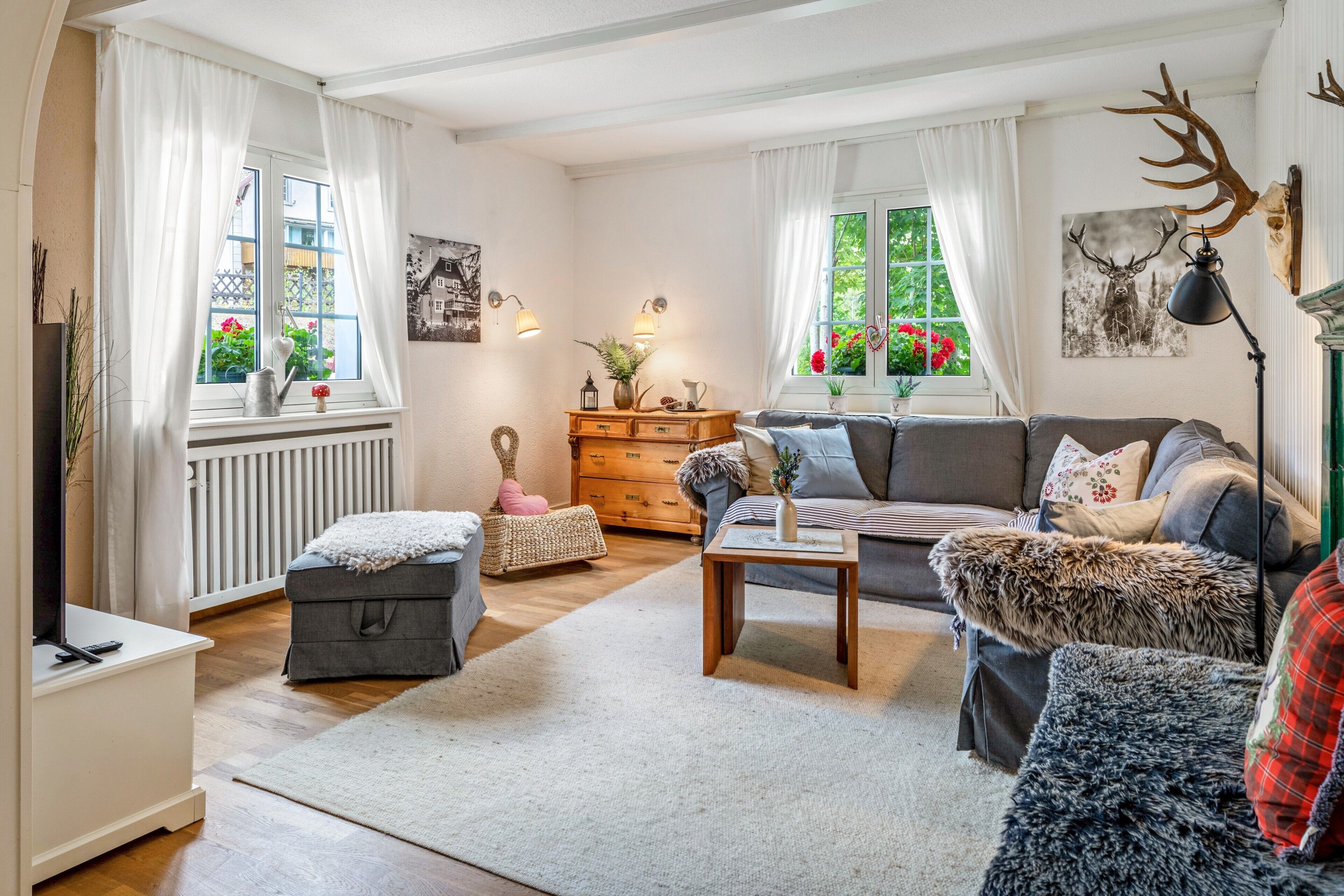 A cozy living room with a sofa, ottoman, and a coffee table. There are windows with curtains, a radiator, and a wall-mounted lamp.