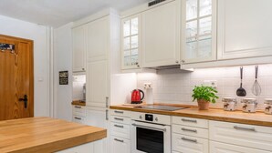 Une cuisine avec des meubles blancs, un plan de travail en bois, une bouilloire rouge, une plante en pot et une horloge.