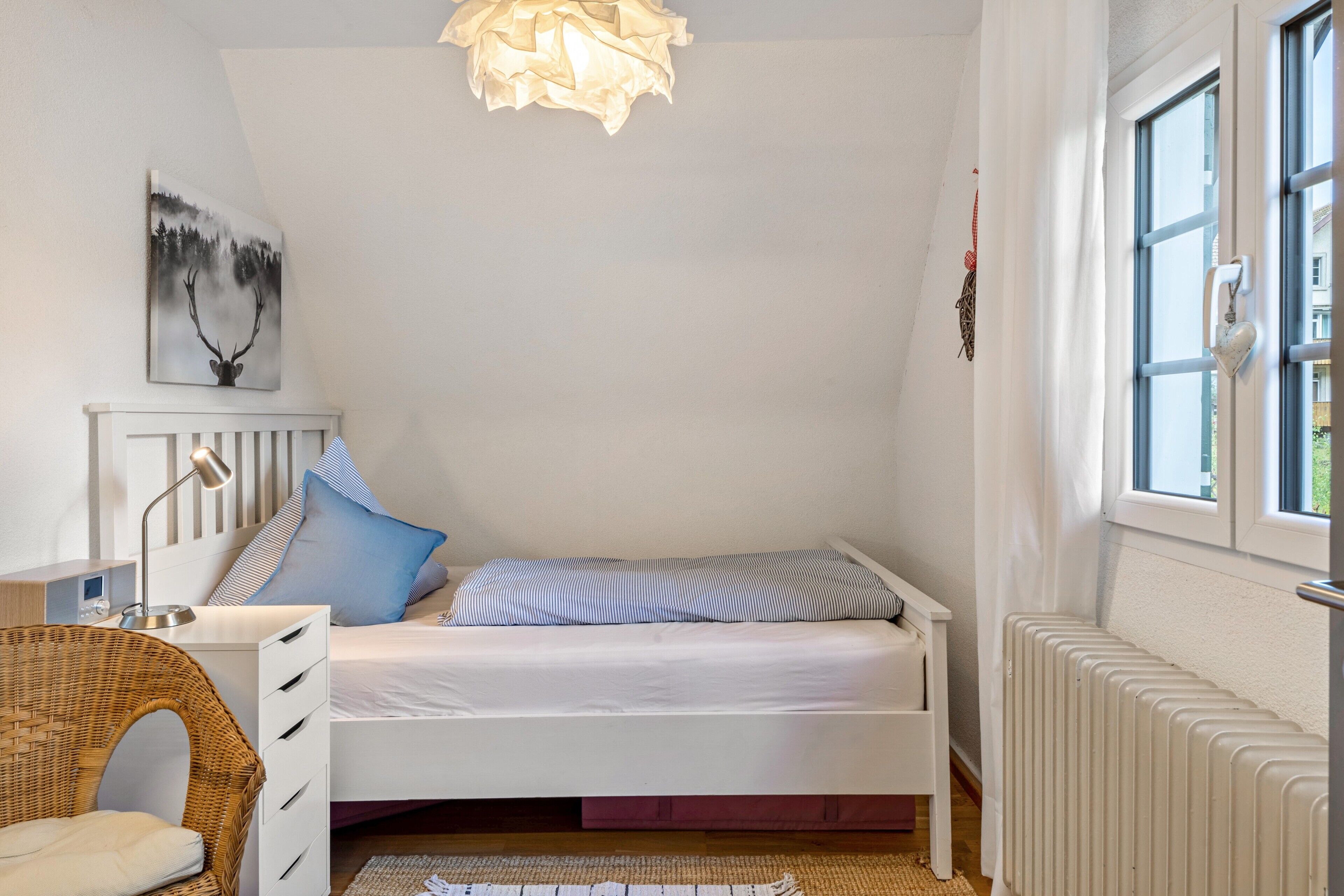 A bedroom with a bed, a bedside table, a lamp, a chair, a radiator, and a window.