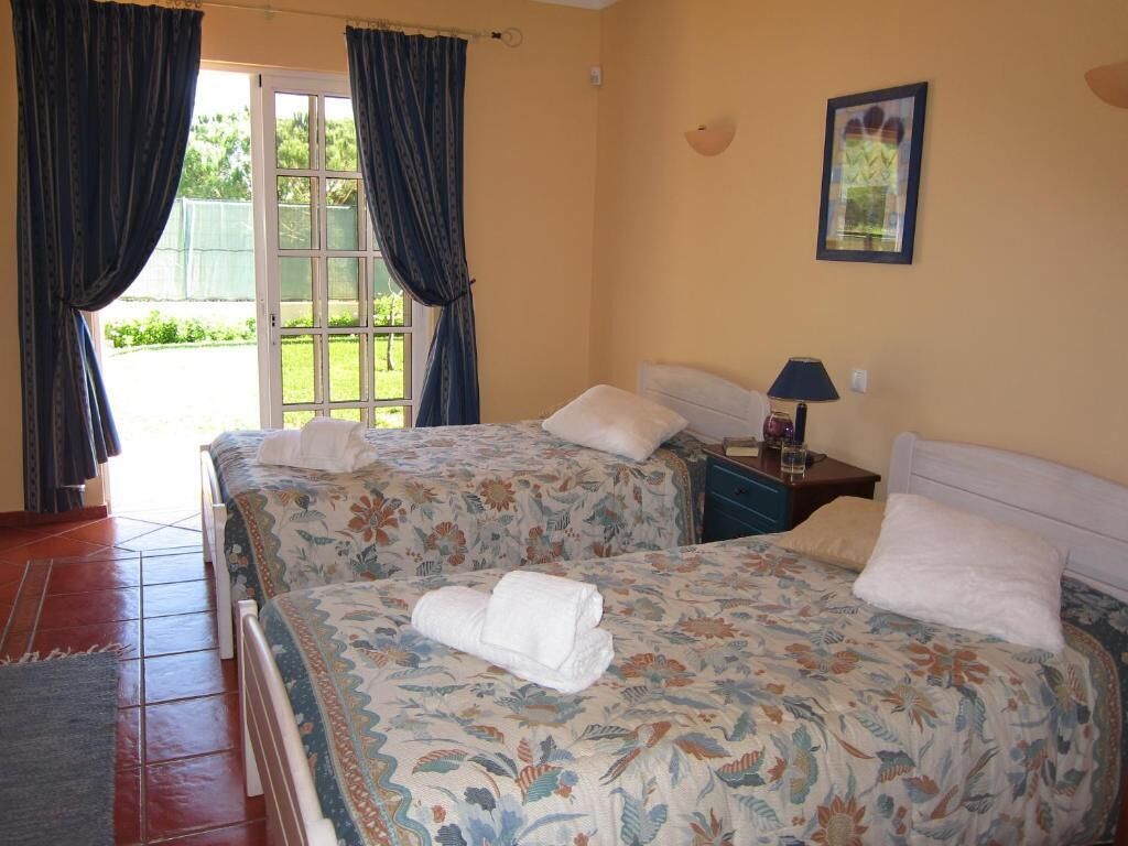 A room with two beds, each with floral bedding and white pillows, a nightstand with a lamp, and a framed picture on the wall.