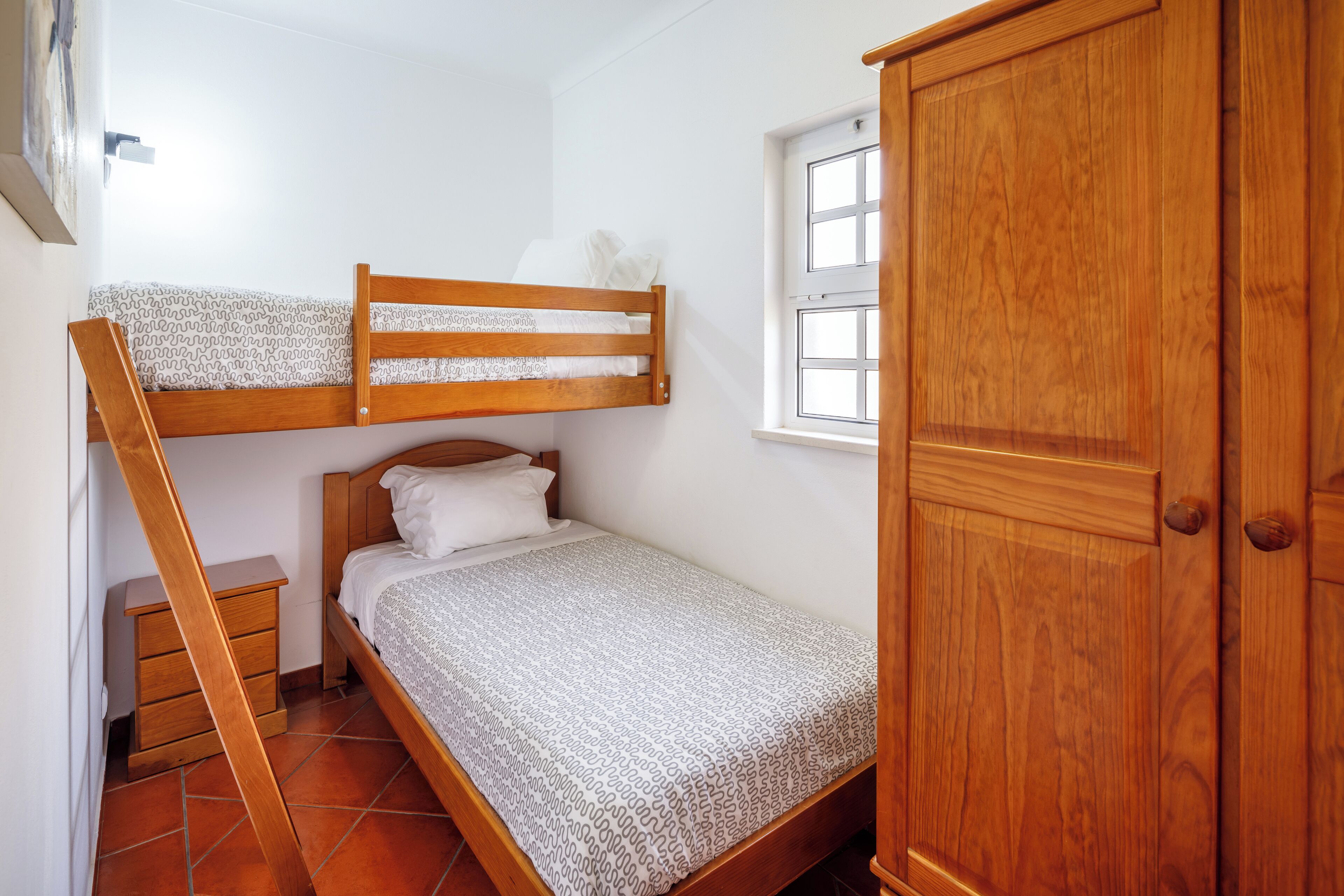 A bedroom with a bunk bed, a single bed, a wooden wardrobe, a bedside table, and a window.