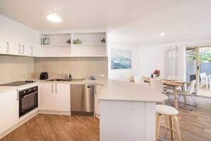 A modern kitchen with white cabinetry, a dishwasher, and a dining area with a table and chairs. - Peaceful Garden Hideaway—Swim, Hike & Unwind (Dunsborough)