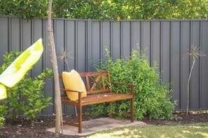 A wooden bench with a cushion, positioned in front of a dark fence and surrounded by greenery. - Peaceful Garden Hideaway—Swim, Hike & Unwind (Dunsborough)