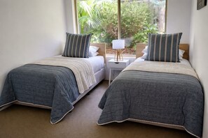 A room with two single beds, a nightstand with a lamp, and a window with a view of greenery. - Peaceful Garden Hideaway—Swim, Hike & Unwind (Dunsborough)