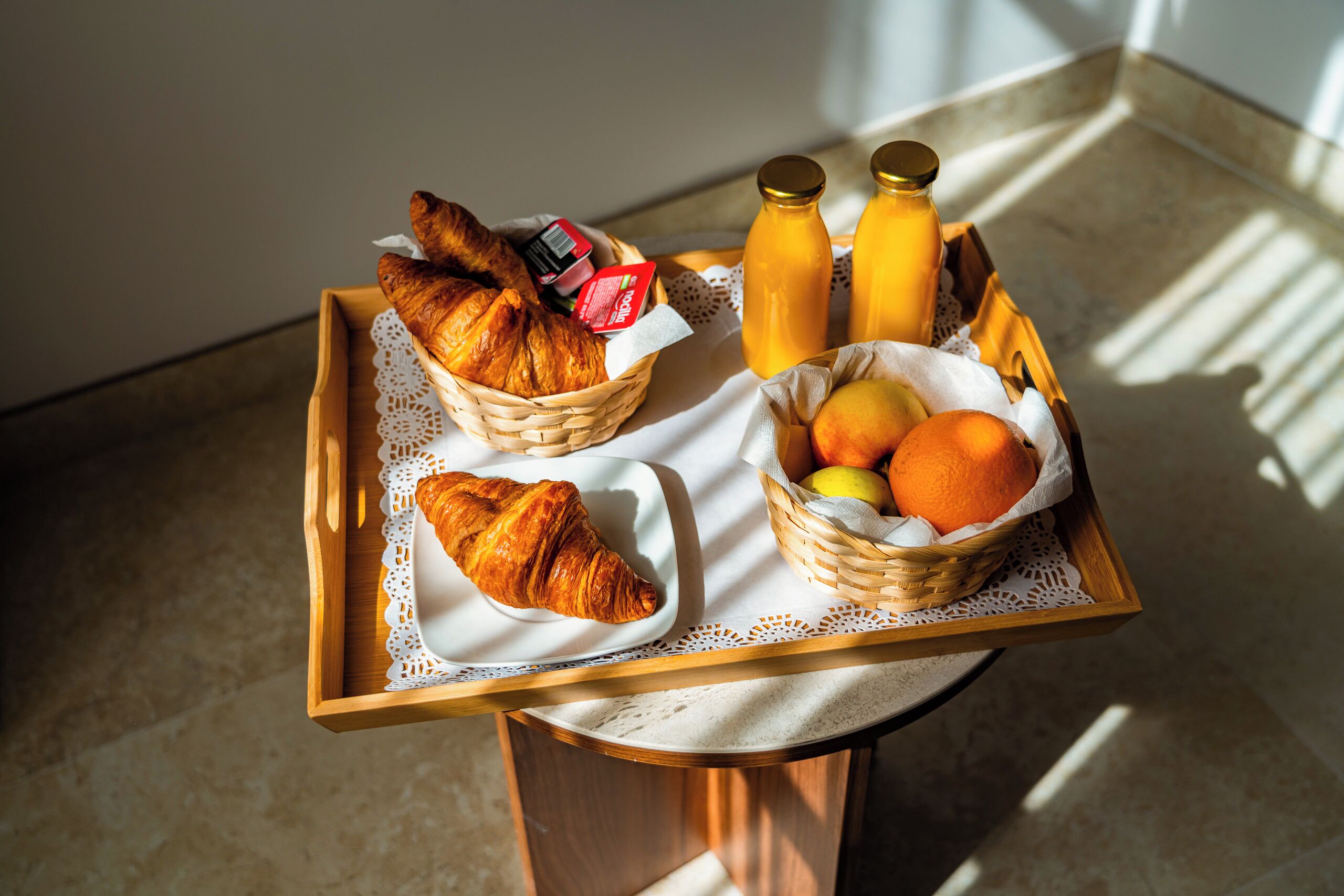 A breakfast tray with croissants, juice bottles, and a fruit basket on a table.