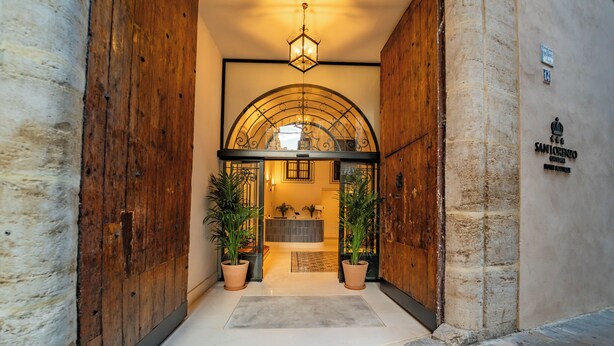 A hotel entrance with a wooden door, a tiled floor, and a reception area visible through an arched window.