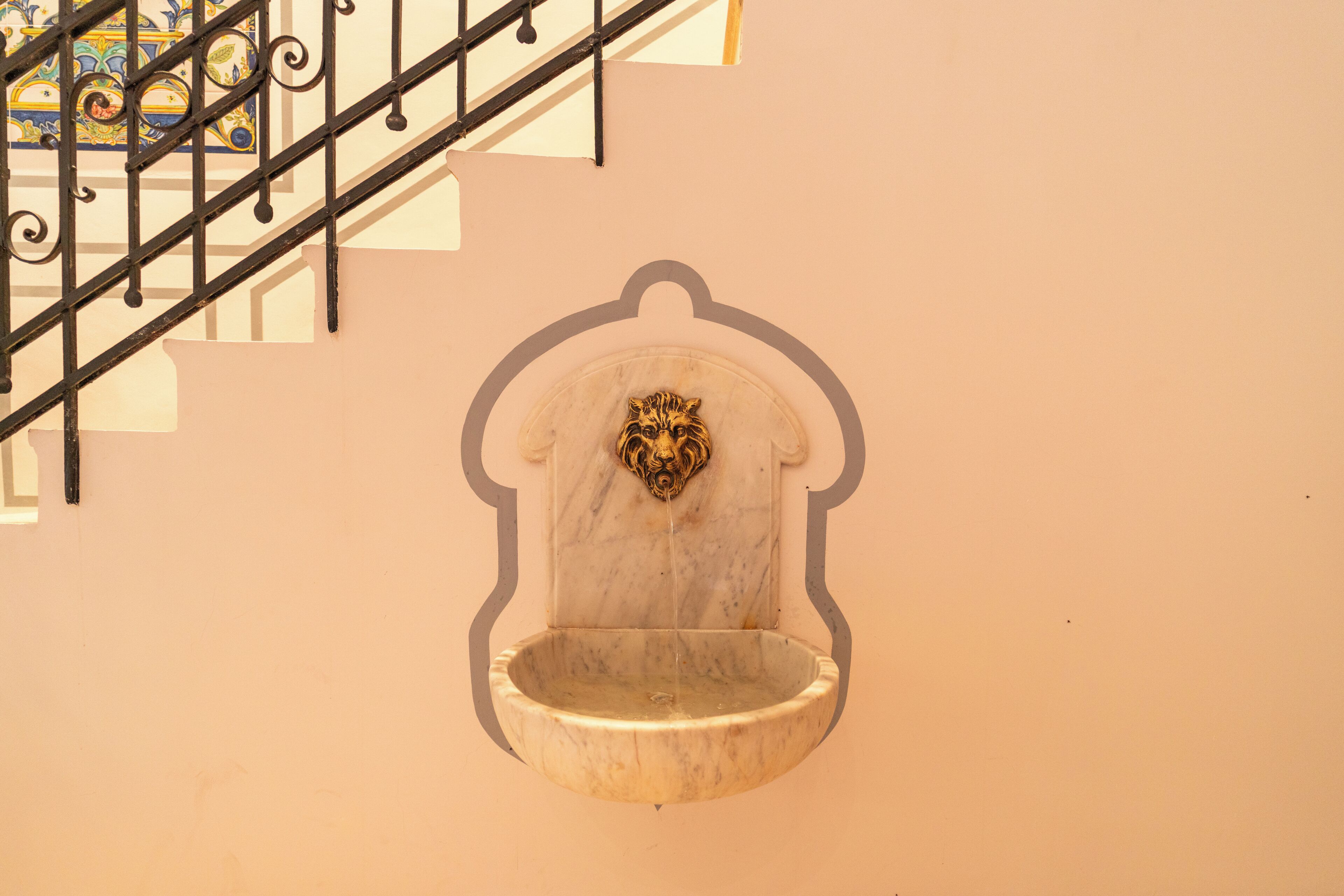 Una fuente de agua empotrada en una pared de mármol con cabeza de león decorativa, una escalera con barandillas de metal negro y un colorido diseño de azulejos en el fondo.