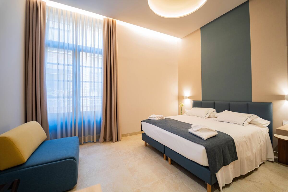 Une chambre d’hôtel moderne avec un lit, un canapé bleu et une grande fenêtre avec des rideaux.