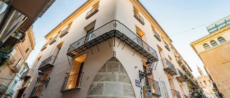 A narrow street with a building featuring a stone archway and multiple balconies with wrought iron railings.