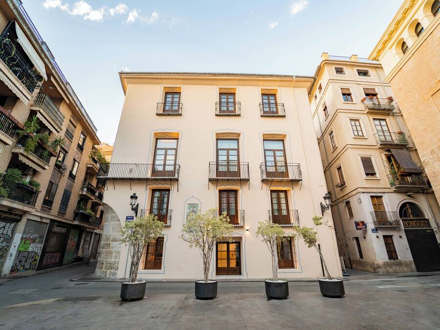 Un edificio de varios pisos con balcones, una entrada central y plantas en macetas en el patio.