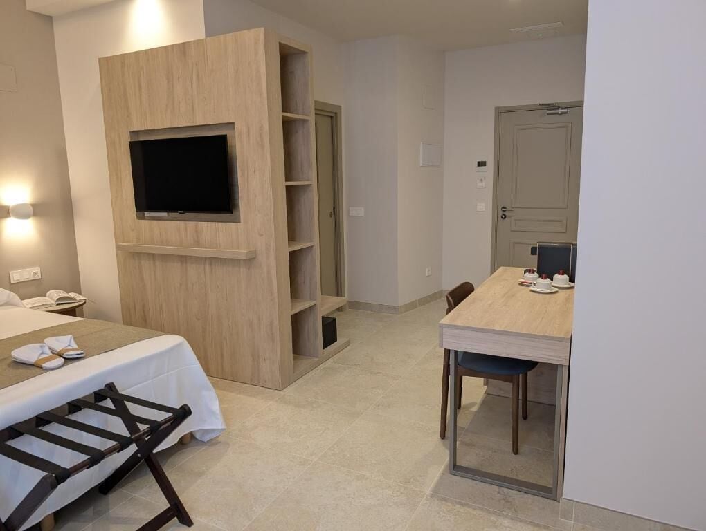 A hotel room with a bed, a TV mounted on a wooden panel, a dining table with chairs, and a door leading to another room.
