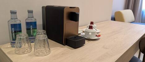 A hotel room with a wooden desk, a coffee machine, two water bottles, and a cup of coffee.