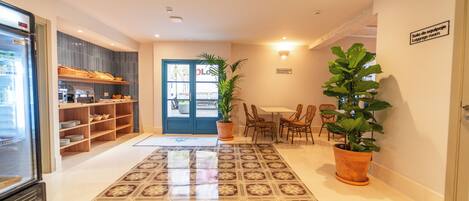 Um hall de hotel com uma mesa de recepção, área de estar com cadeiras, uma grande planta em vaso e piso ladrilhado.