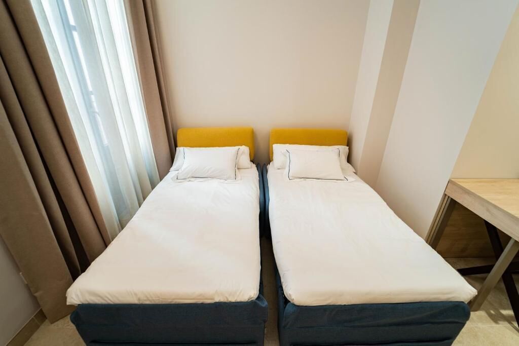 A small, single-room accommodation with two single beds, a wooden desk, and a window with curtains.