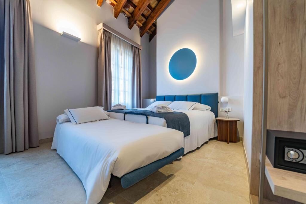 A hotel room with two beds, a circular light fixture, a wooden ceiling, and a small bedside table.