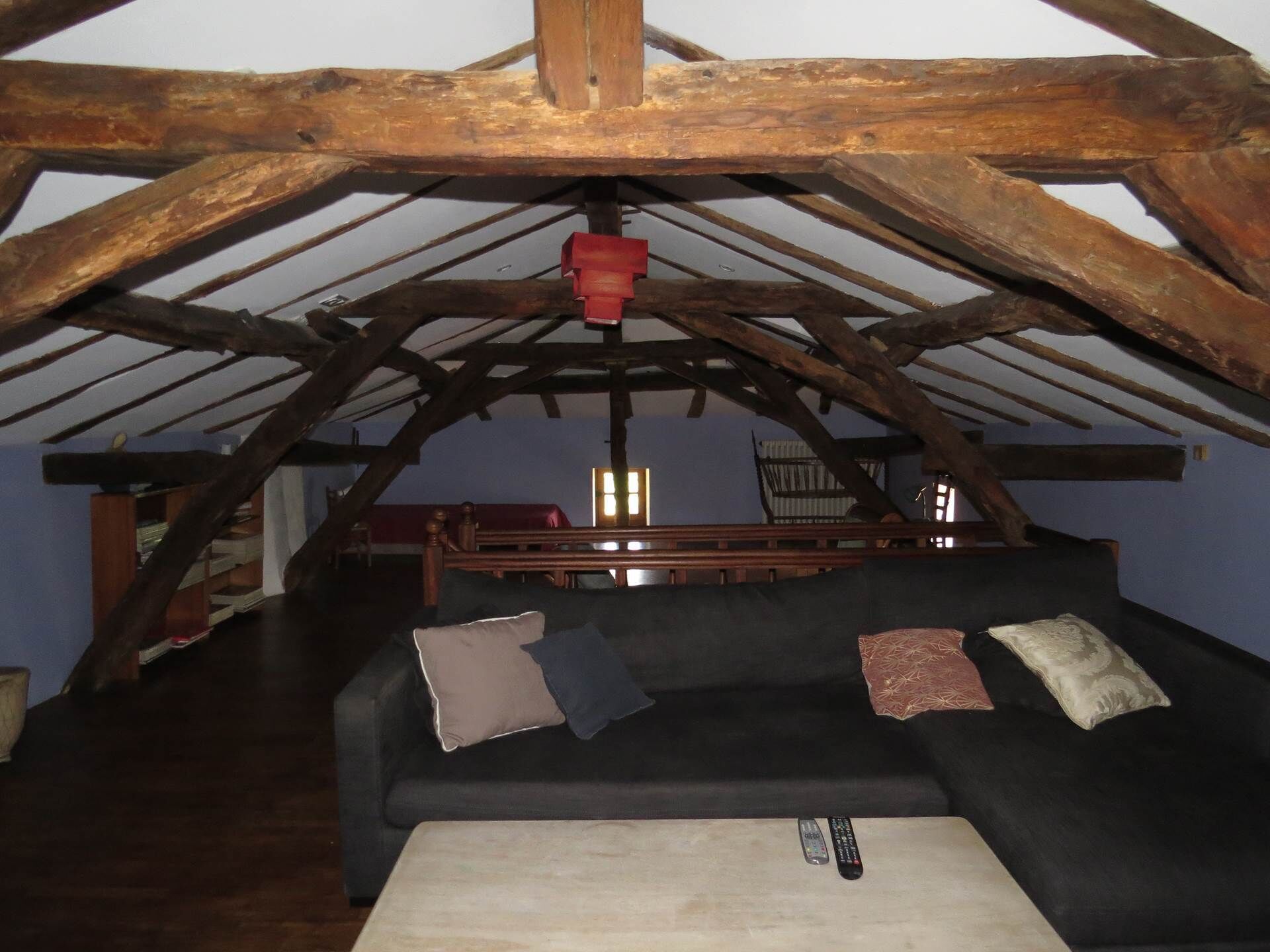 Ein gemütliches Dachzimmer mit Holzbalkendecke und sichtbaren Holzbalken, einer schwarzen Couch mit Kissen und einem kleinen Tisch mit Fernbedienung.
