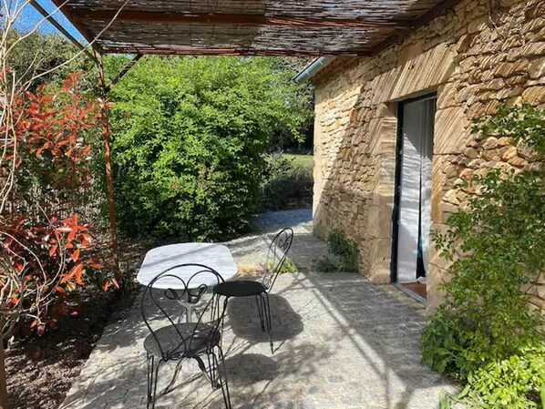Un bâtiment en pierre doté d’une porte, d’un patio couvert avec une table et des chaises, et entouré de verdure.