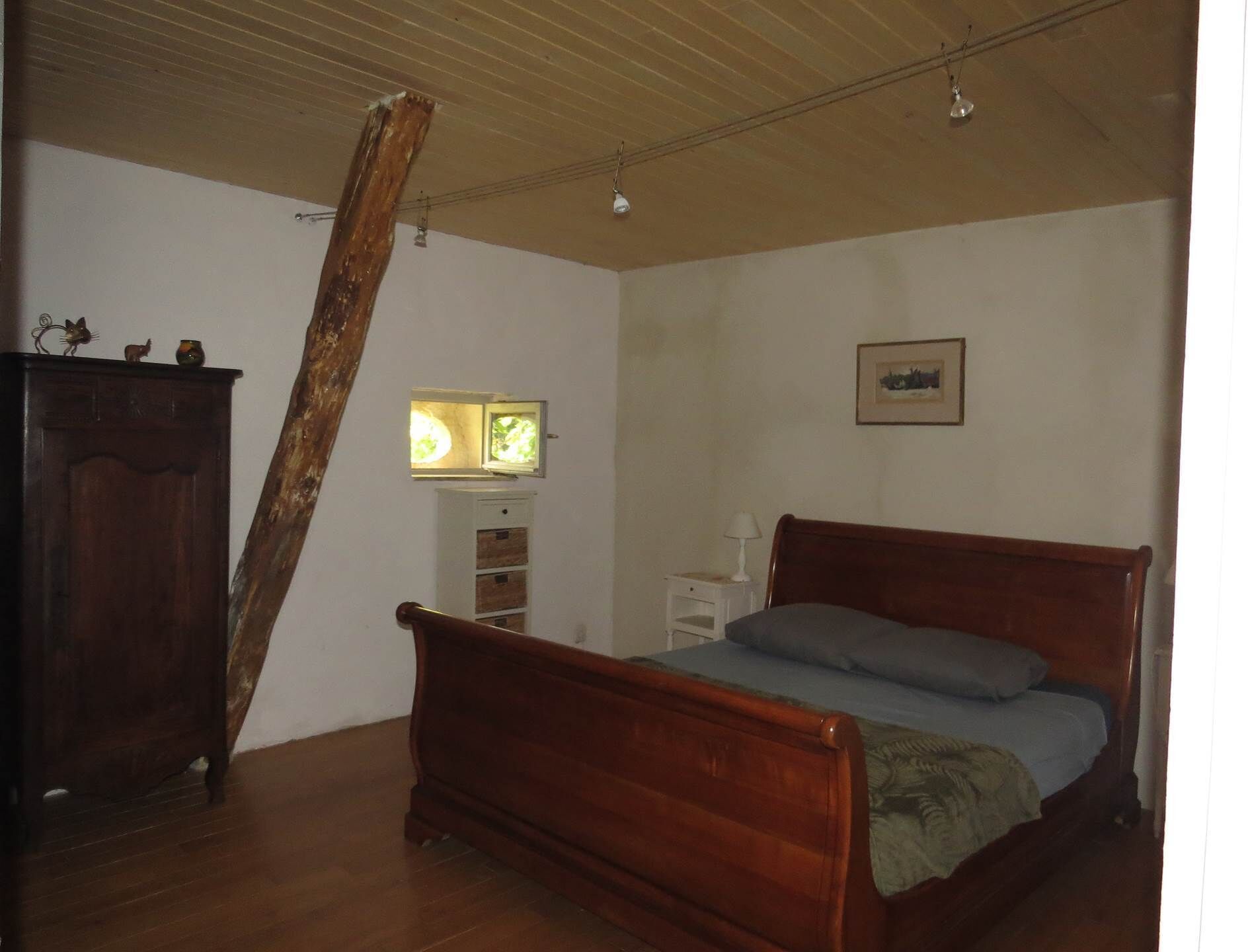 Ein Schlafzimmer mit einem hölzernen Bett, einem Nachttisch, einer Kommode und einem Fenster mit Blick auf Grünfläche.