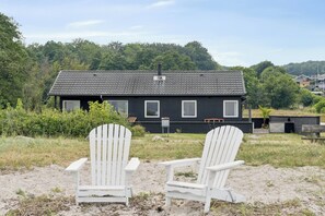 A two-story house with a dark exterior and a tiled roof, surrounded by greenery and a sandy area with two white Adirondack chairs. - Renovated beach house with unique location (Bjert)