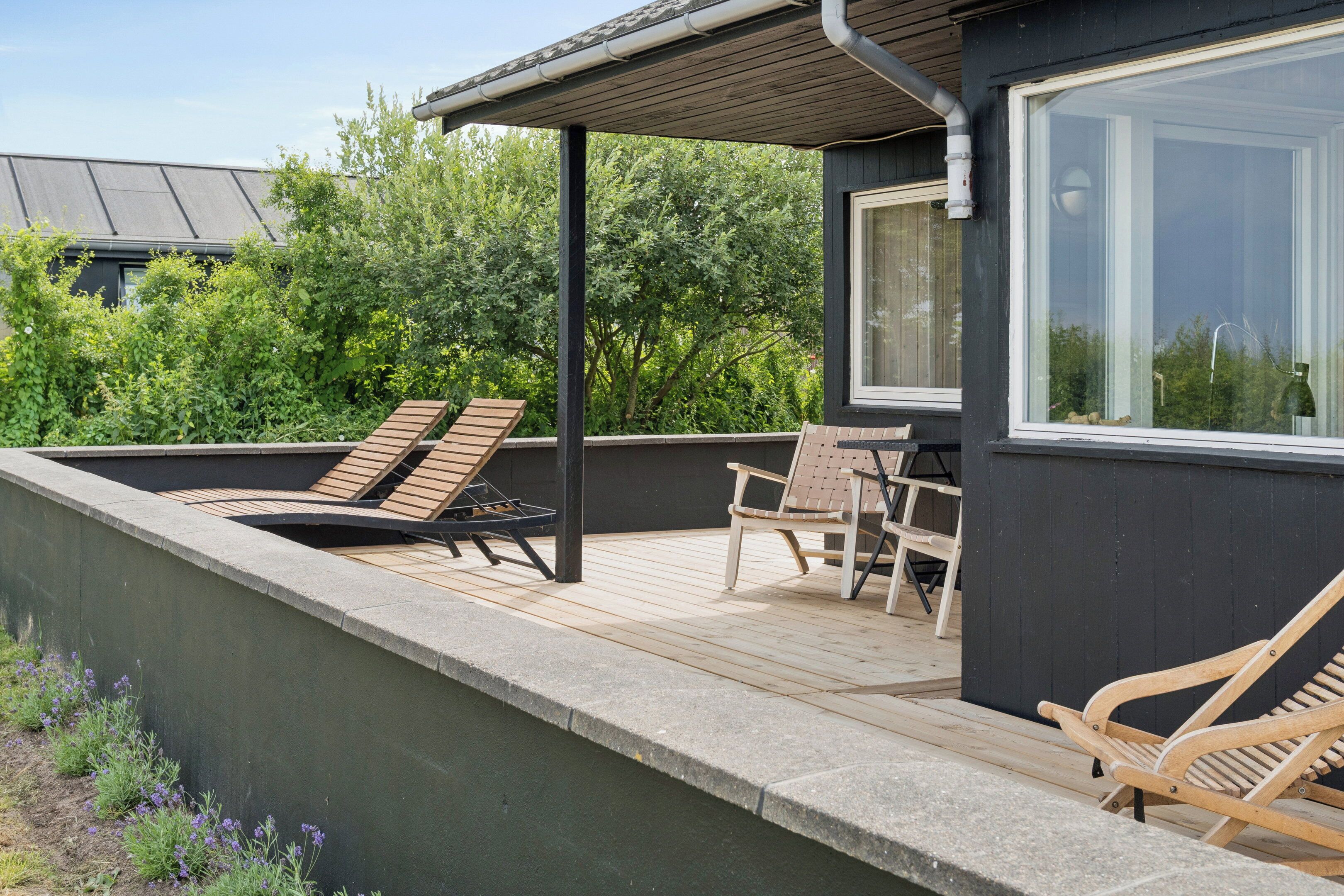 A rooftop terrace with a wooden deck, two lounge chairs, a table, and a window overlooking greenery.
