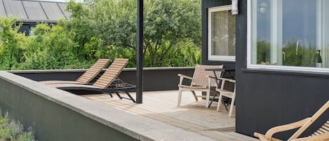 A rooftop terrace with a wooden deck, two lounge chairs, a table, and a window overlooking greenery.