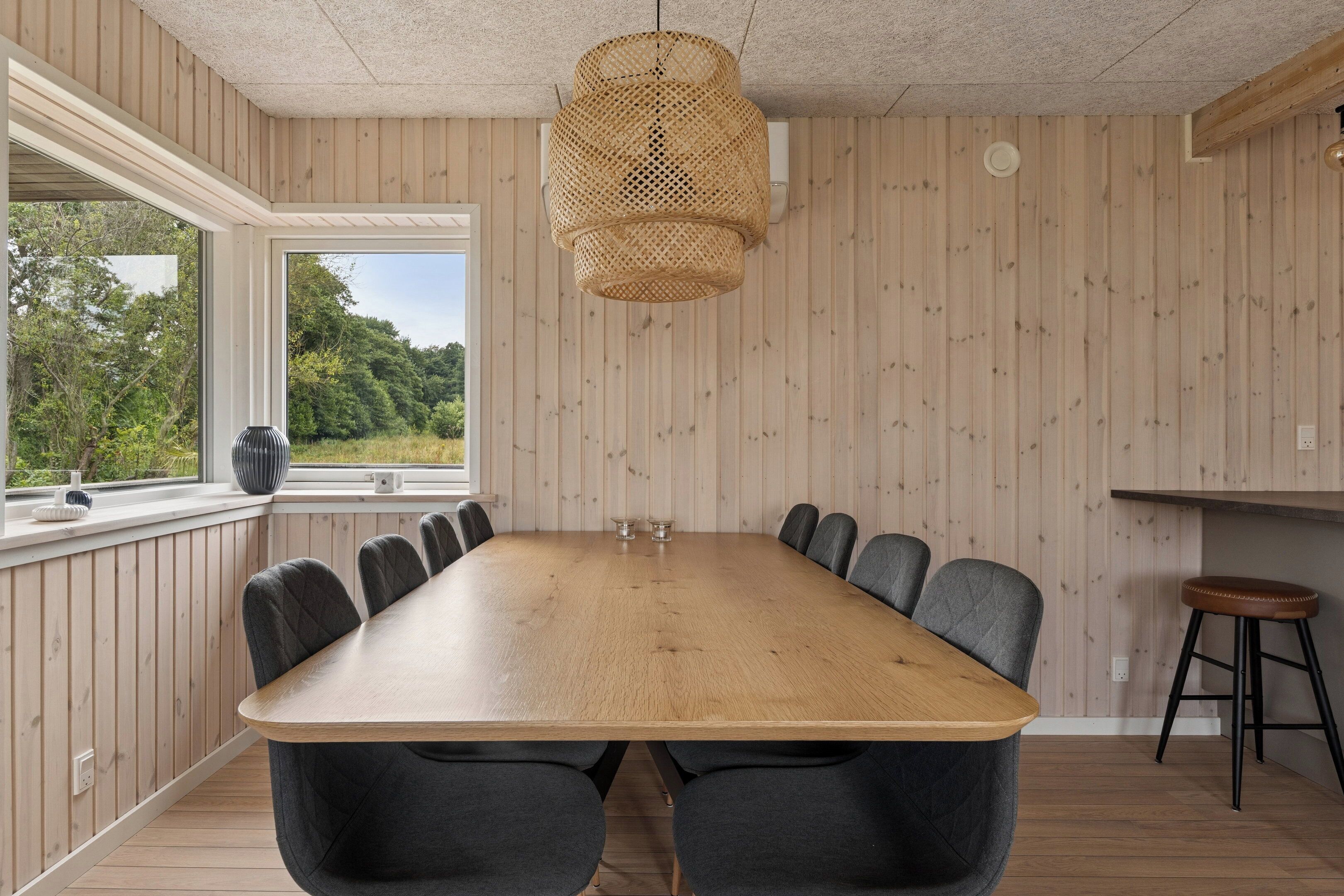 A conference room with a long wooden table, dark chairs, a woven pendant light, and a window with a view of greenery.