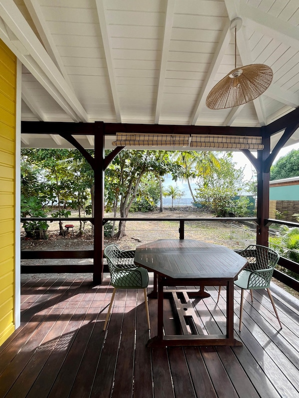 A covered outdoor area with a wooden table and chairs, a hanging light fixture, and a view of greenery and a body of water.