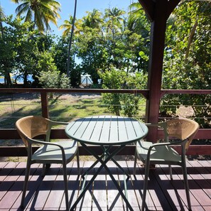 Un patio exterior con una mesa redonda y dos sillas de mimbre, rodeado por una terraza de madera y vistas a árboles tropicales.