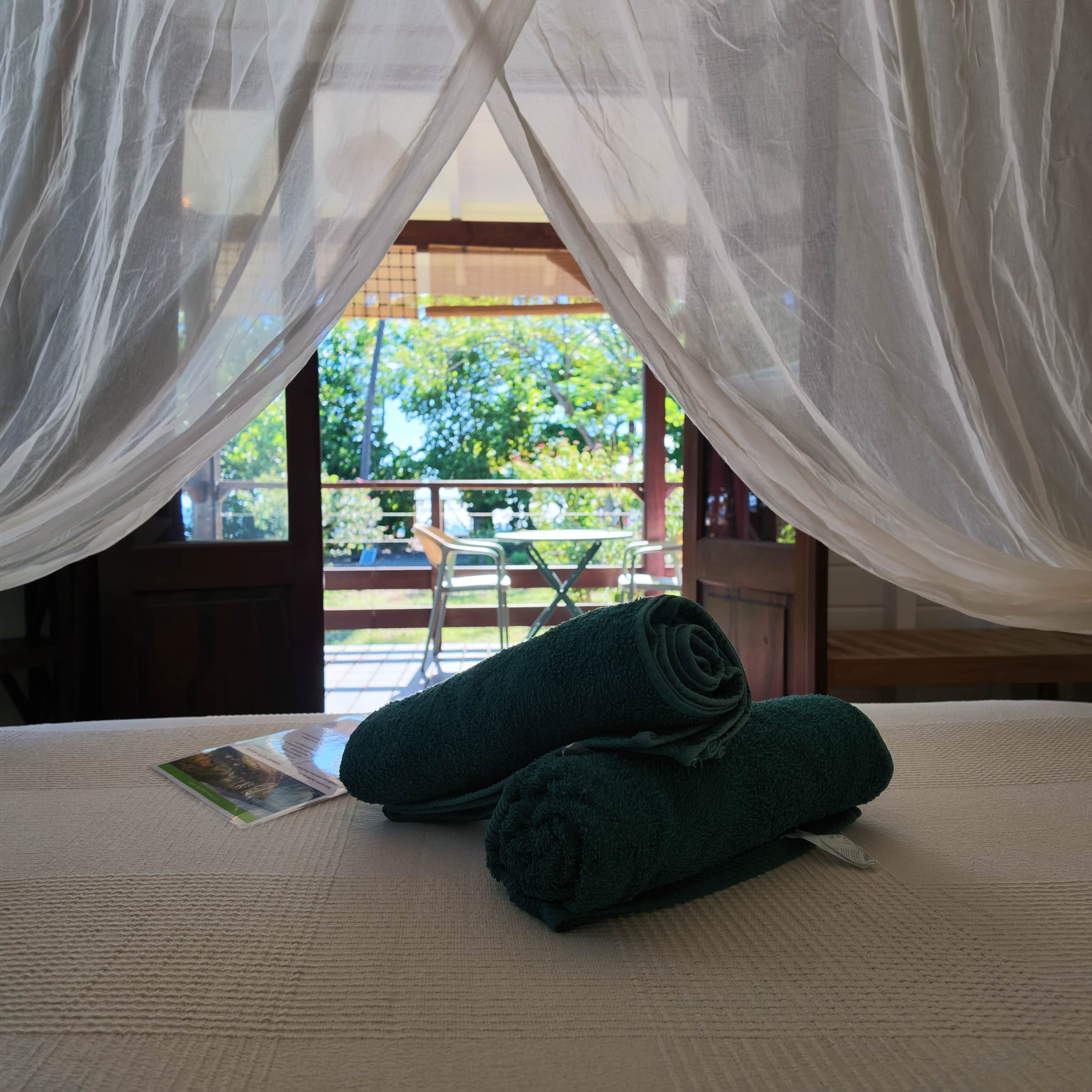 A bed with a canopy, a pair of rolled towels, and a view of an outdoor area with a table and chairs.