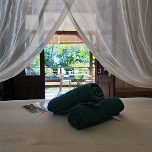 A bed with a canopy, a pair of rolled towels, and a view of an outdoor area with a table and chairs. - Beach Bungalow: Coulirou (Le Carbet)