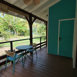 A covered outdoor area with a table and chairs, a teal door, and a view of greenery. - Beach Bungalow: Thazard (Le Carbet)