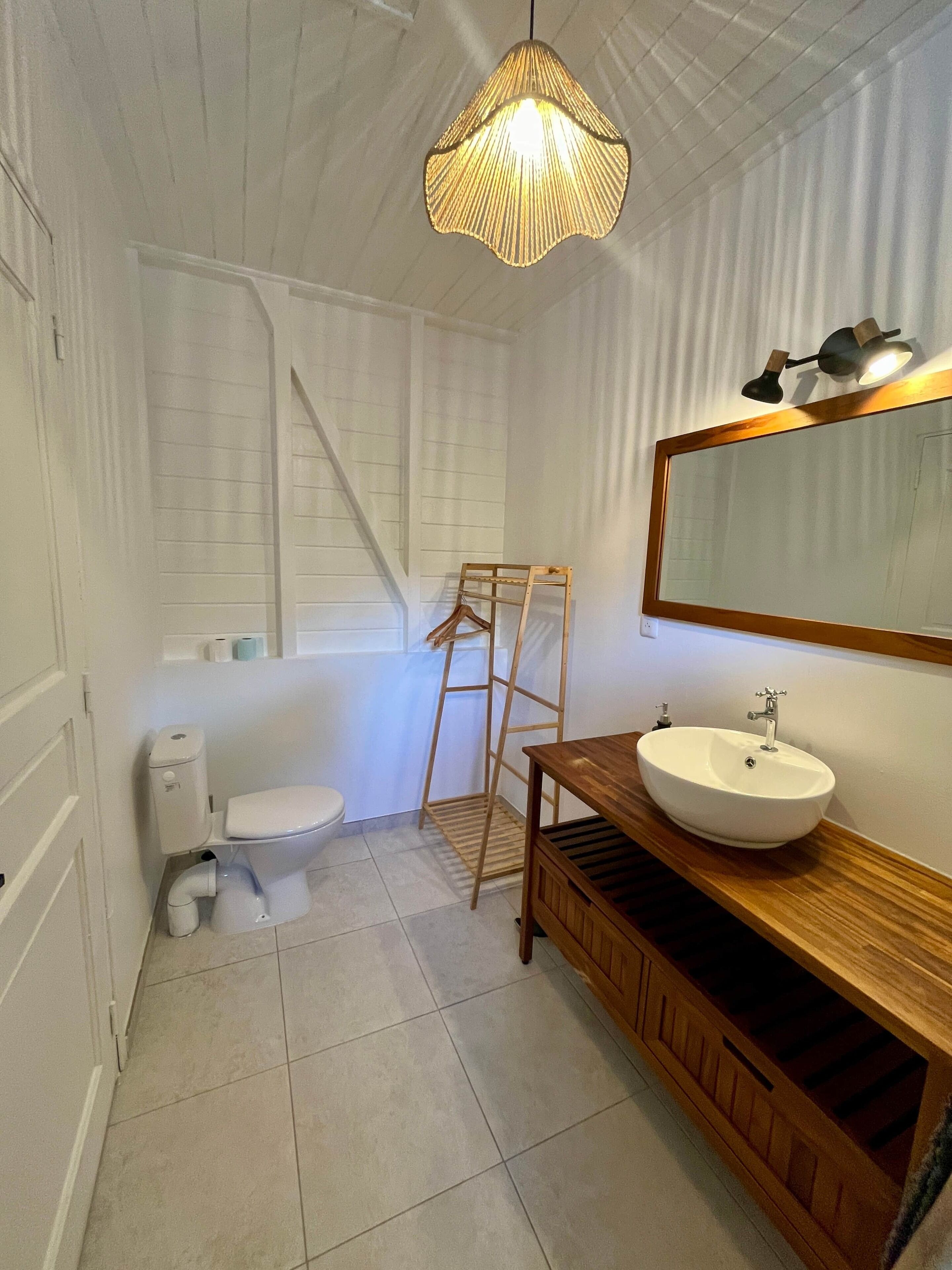 Un baño con inodoro, lavamanos, tocador de madera, espejo y una lámpara colgante.