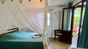 A room with a bed, a wooden chair, a table with a book, and a window with curtains. - Beach bungalow: Balaou (Le Carbet)